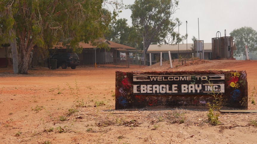 A sign that says "Welcome to Beagle Bay" in front of a dusty outback settlement.