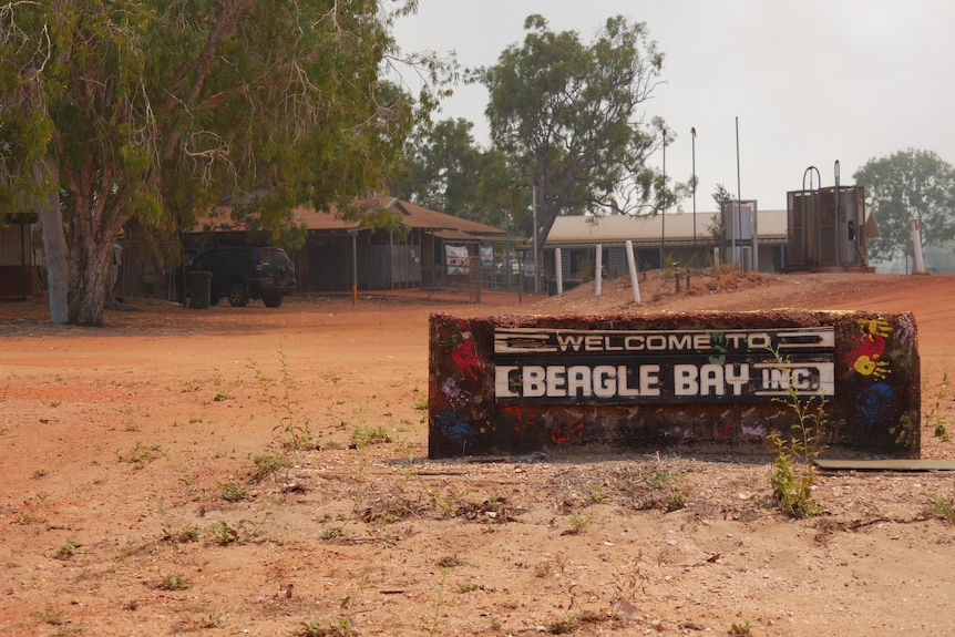 A sign that says "Welcome to Beagle Bay" in front of a dusty outback settlement.