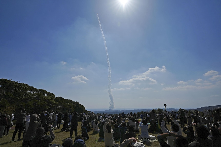 Crowds watch a rocket climb into the sky.