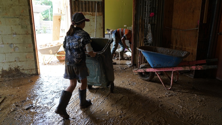 Young girl wheels a green bin across a muddy floor, as a man in orange high vis picks up debris in background.