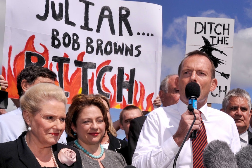 Tony Abbott stands with a microphone in front of a crowd holding signs. One says 'ditch the witch' 'Juliar Bob Brown's bitch'