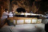 Archaeologists remove the cover of an intact sarcophagus inside the tomb. There is a body wrapped in cloth inside