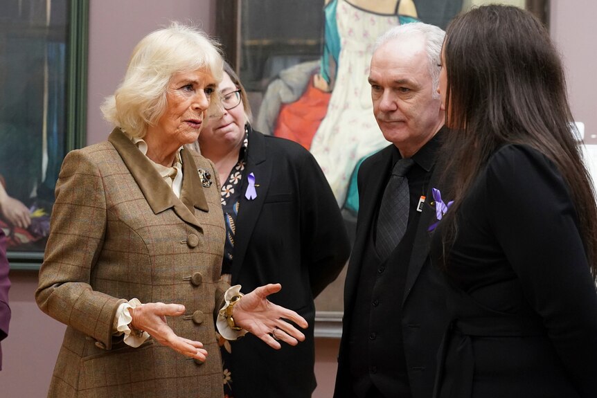 A medium shot of Queen Camilla speaking to three people and gesturing with her hands in an art gallery.