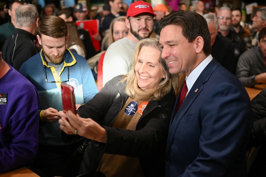 Ron DeSantis smiles and poses for a selfie with a woman in amongst a crowd of people.