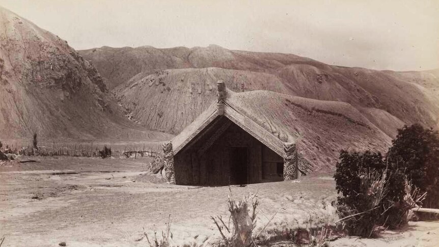A black and white image shows a Hinemihi meeting house in the middle of ash-covered valley with mountains along the horizon.