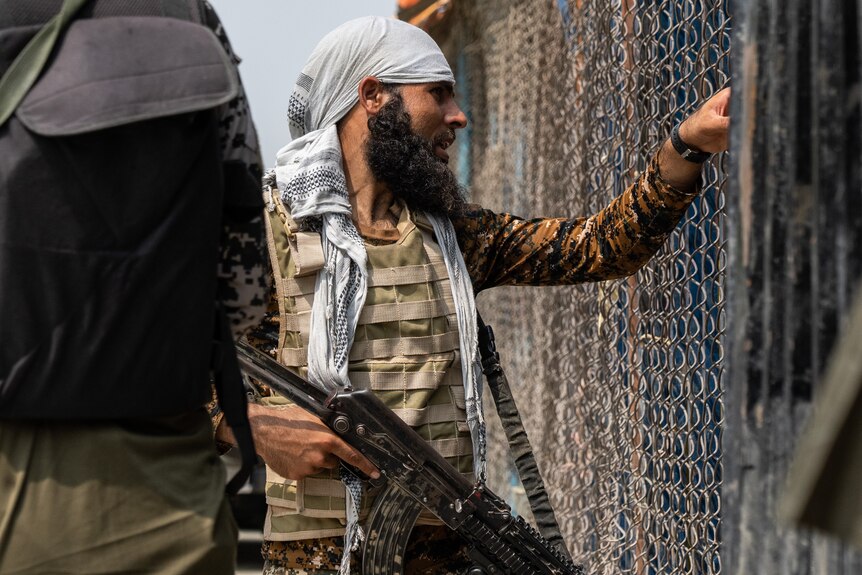 A bearded man with a camo flack jacket and gun looks through a wire fence