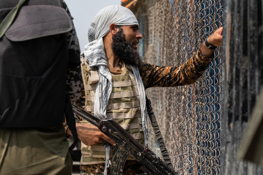 A bearded man with a camo flack jacket and gun looks through a wire fence