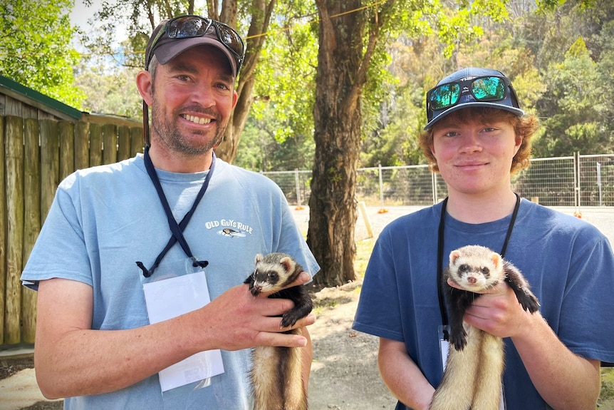 A man and a boy hold up a ferret in their hands.