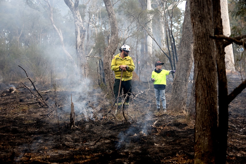 A man with a rake and a yellow shirt standing in burnt bushland watches on as a fire smoulders