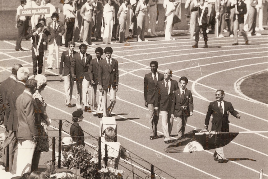 Men in sporting blazers walk on a running track in a parade.
