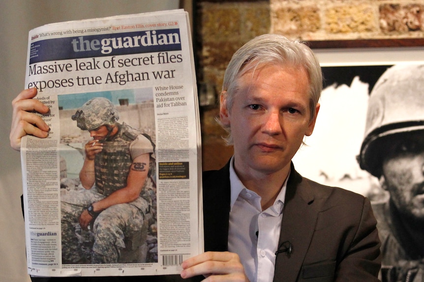 Assange, looking younger, holds up a newspaper reading a massive leak of secret files revealing the real Afghan war.