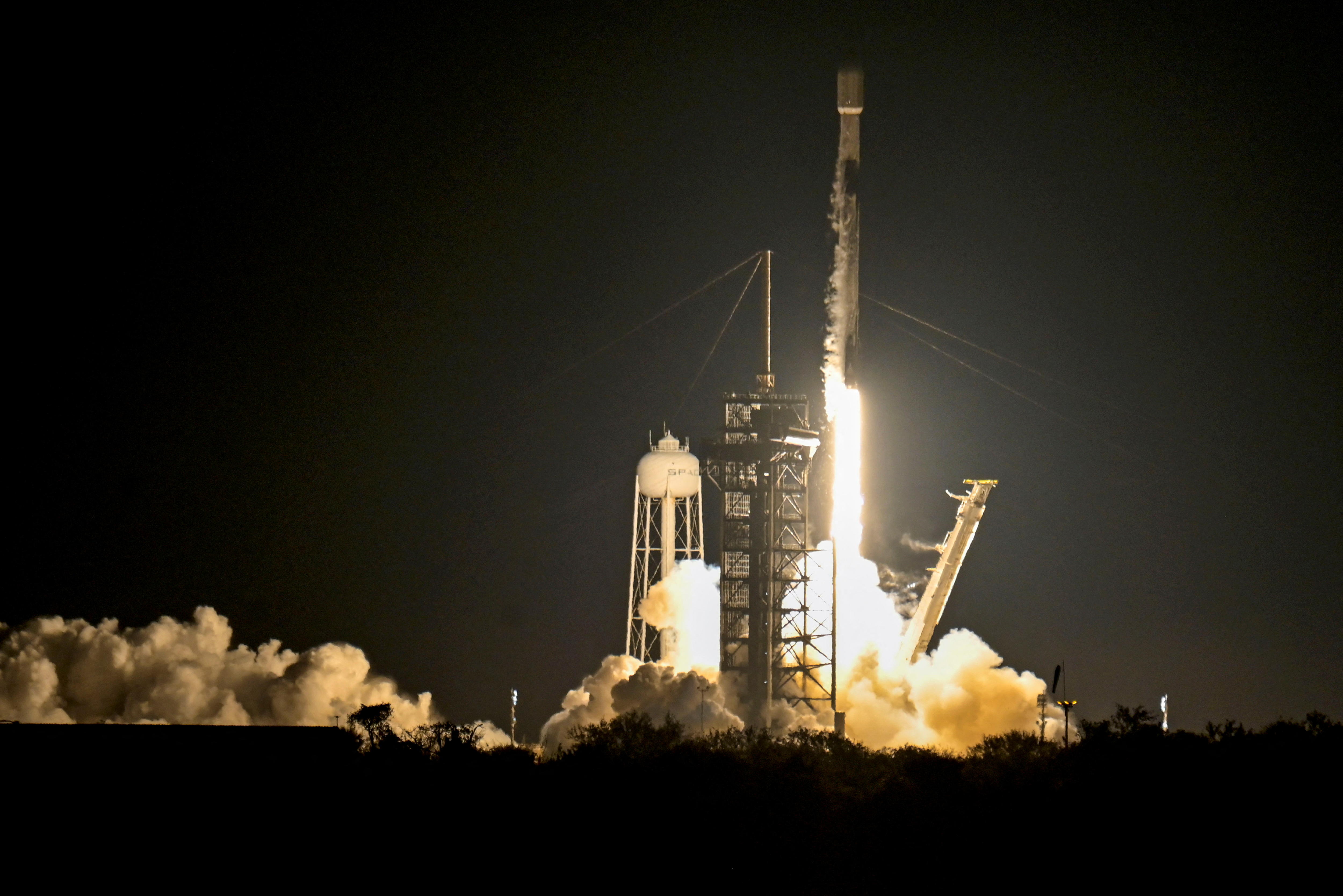 A SpacX rocket blasting off from a launchpad at night, surrounded by bright yellow flames and smoke plumes