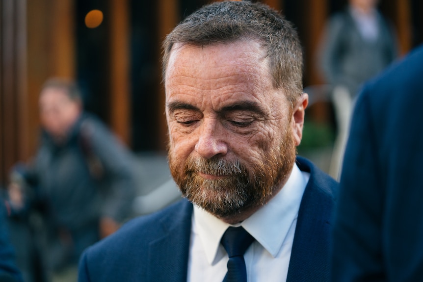 A close up of Michael wearing a navy blue suit and white shirt, with a slight frown, walking outside with his eyes down.