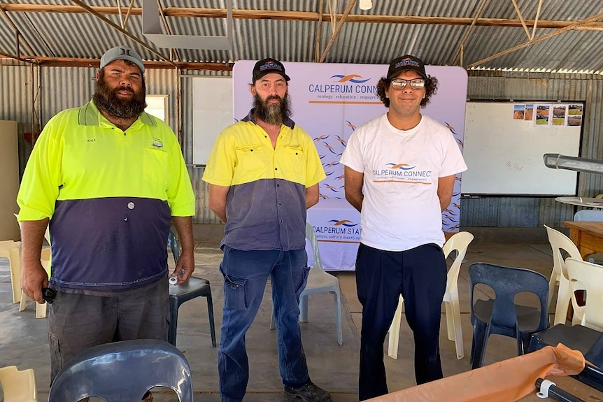 Three men are standing in a shed. Two are wearing hi-vis and one is in a white t-shirt.