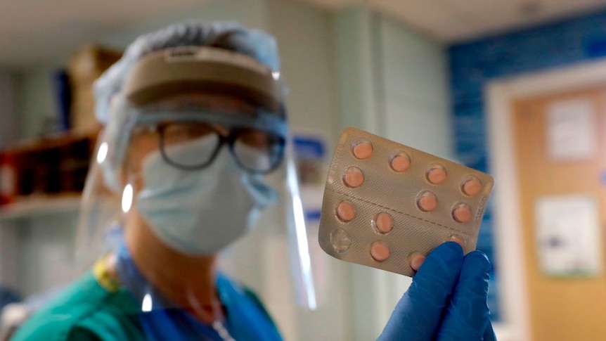 A female nurse dressed in protective mask and gown holds a packet of pills.