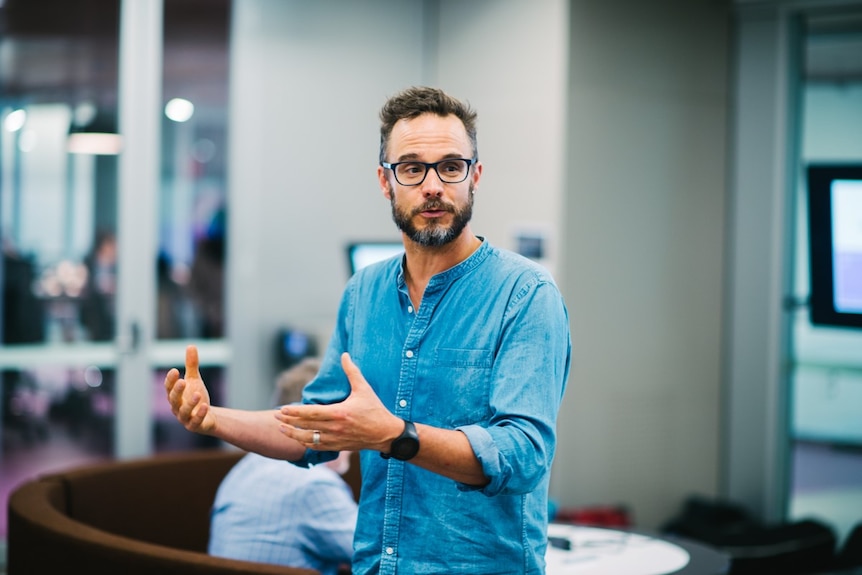 A man inside a well-lit room, wearing a blue shirt and glasses looking away from the camera and using his hands to speak.