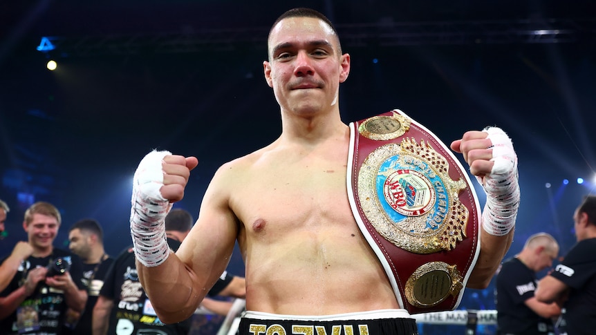 Boxer Tim Tszyu smiles at the camera, with fists clenched and a world title belt slun over his shoulder.