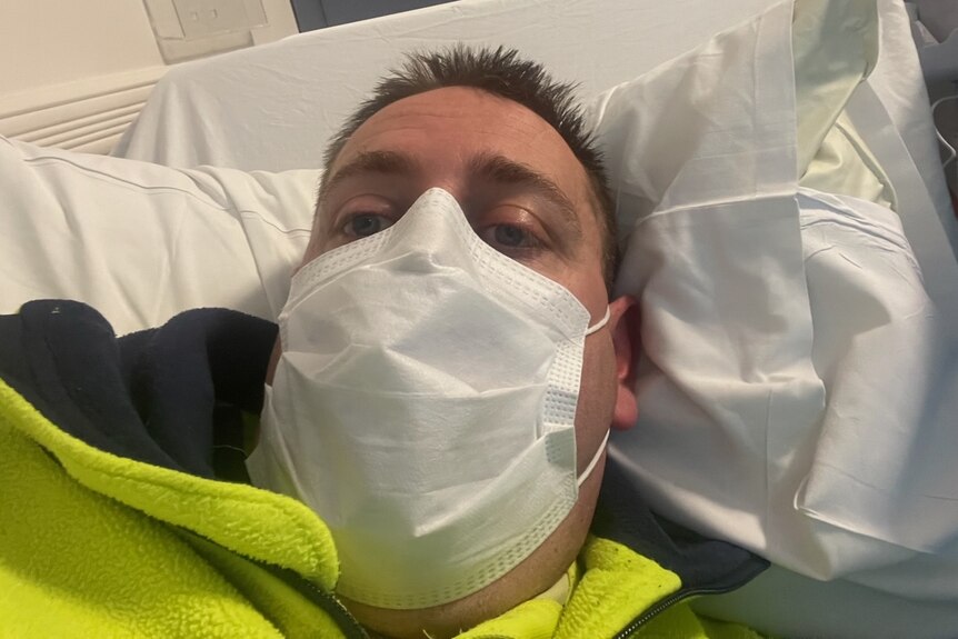 James Mancey wearing a high vis jumper laying in a hospital bed with a mask. 