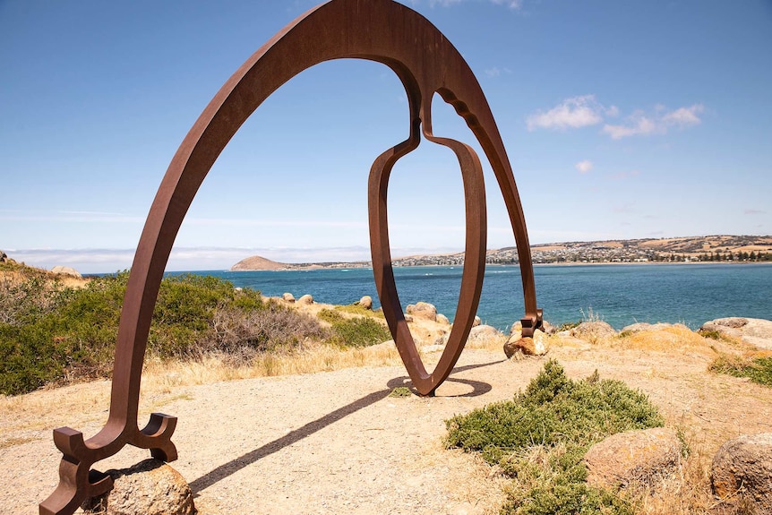 A sculpture resembling a steel arch sits on rocks with the Victor Habor shoreline in the distance