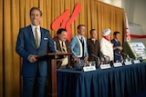 A man at podium smiles at the camera in front of five more men behind a long table.