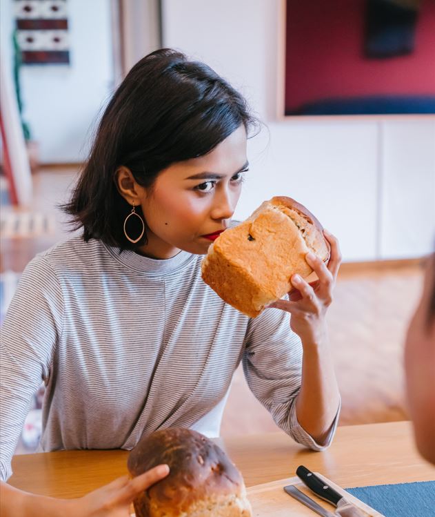 Woman sits at a table and holds a loaf of bread to her nose as if smelling it, and stares ahead.