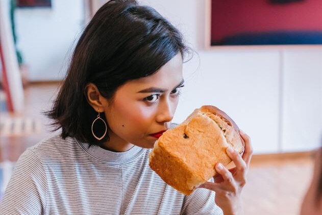 Woman sits at a table and holds a loaf of bread to her nose as if smelling it, and stares ahead.