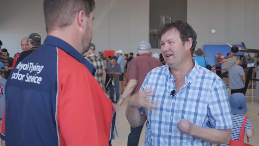 Two men talk to each other; one wears a Royal Flying Doctor Service shirt; the other is missing a hand.