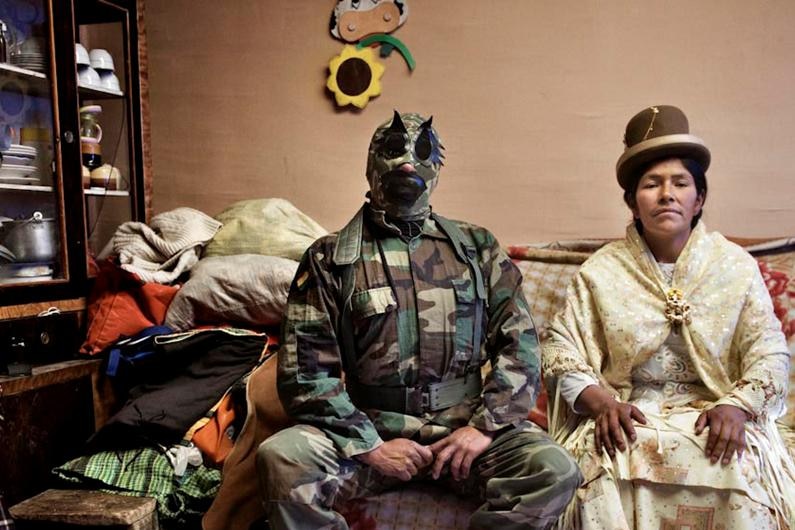 A masked Bolivian wrestler sits in his home, with his wife by his side.