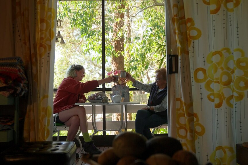 Sally, right, and Eden, left, are seen sitting outside cheersing on a verandah in an image taken from inside framed by curtains