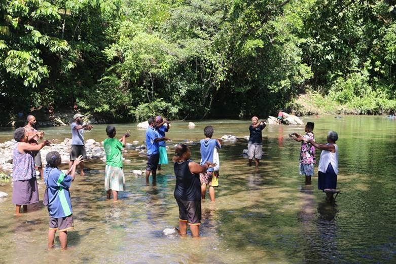 A group of men and women stand in a lake with their hands outstretched.