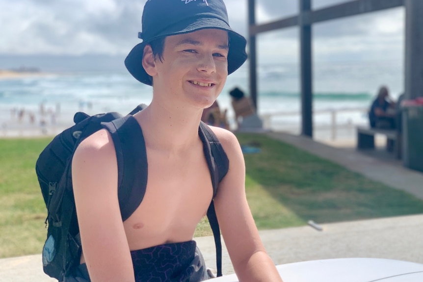 A boy at the beach with a surfboard, smiling.