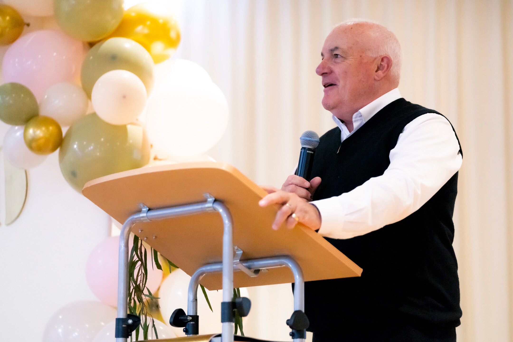 Bald man, wears white shirt, black sleeveless jumper, speaks from podium with gold, white, pink balloons t, holds microphone.