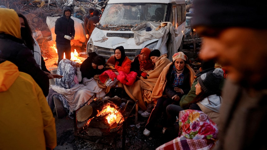 People sit around a fire next to rubble and damages near the site of a collapsed building.