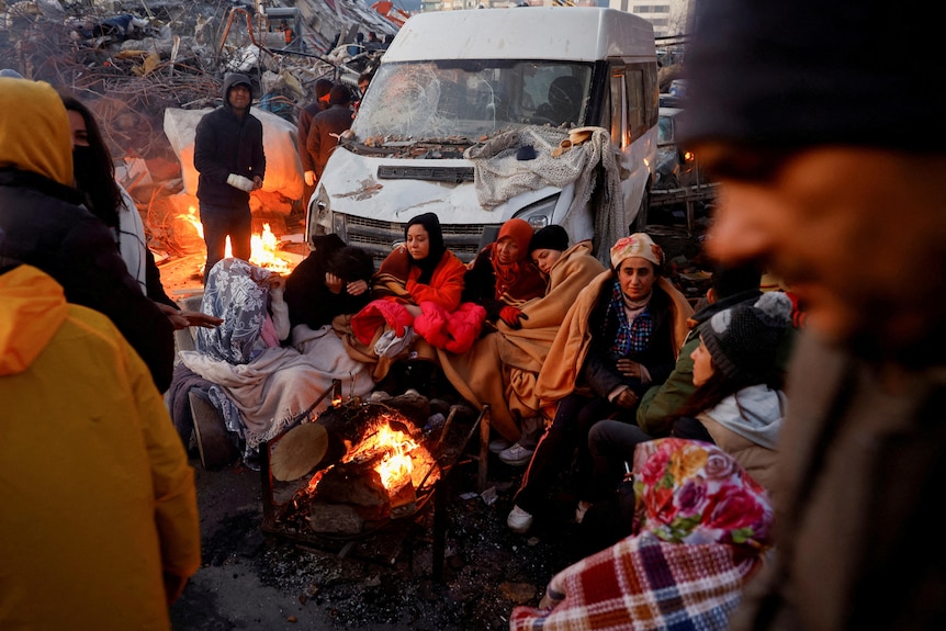 People sit around a fire next to rubble and damages near the site of a collapsed building.