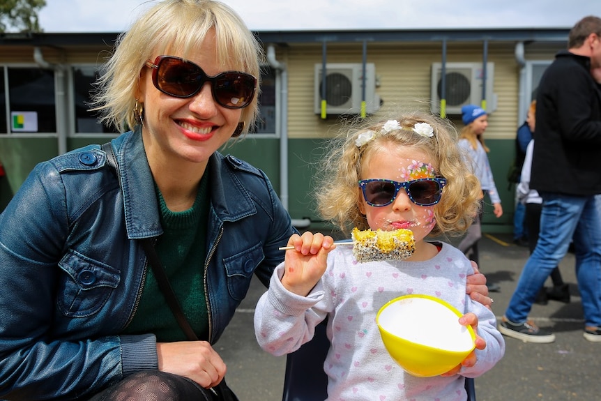 A mum smiles at the camera with her daughter next to her, who is eating corn on a stick with a reusable bowl.