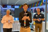Two men and a woman standing in front of an IT store holding mobile phones
