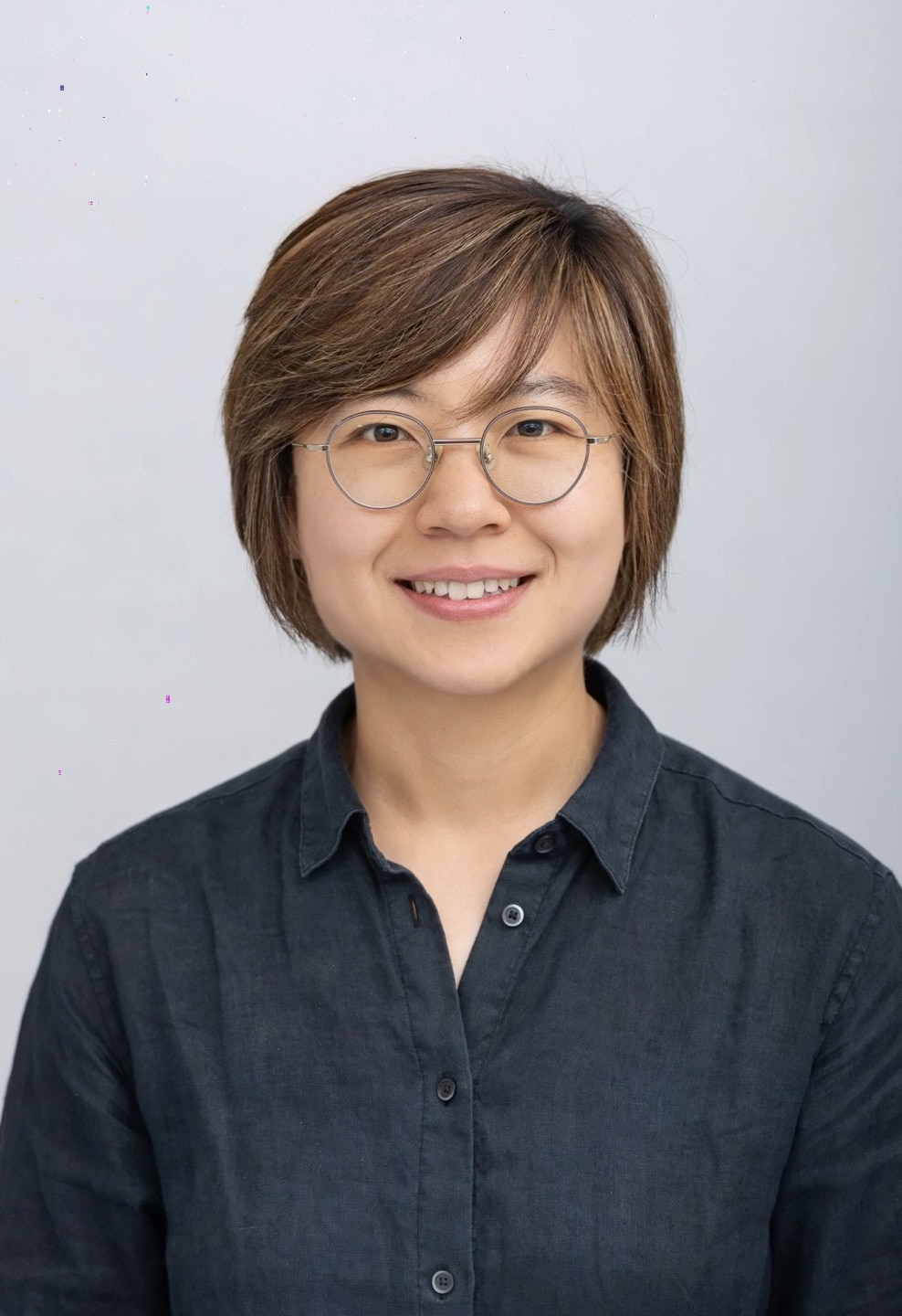 Un retrato profesional de una joven sonriente, Mirim Shin, con gafas y de pie sobre un fondo gris.