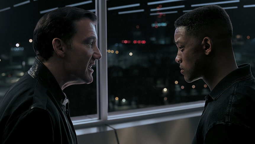 Clive Owen (left) faces Will Smith (right) and yells towards him in front of glass windows at night time.
