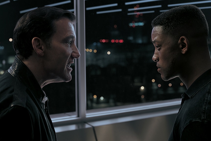 Clive Owen (left) faces Will Smith (right) and yells towards him in front of glass windows at night time.