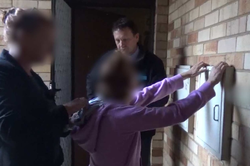 A woman with her hands up against a wall and two police officers surrounding her.