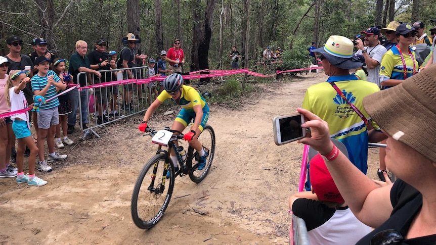 A female mountain bike rider in Australian colours heads uphill past cheering fans.