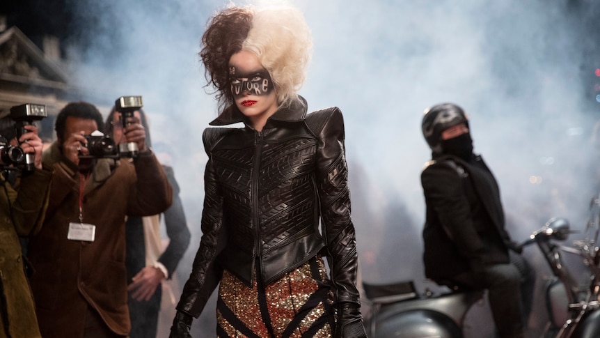 Film still of Emma Stone as Cruella on a red carpet wearing makeup that says The Future, with man on motorcycle behind her