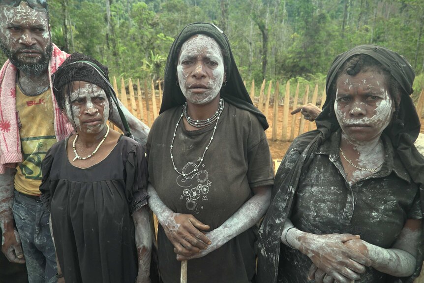 Three women and one man stand in a row with their faces painted white, mourning their lost relatives