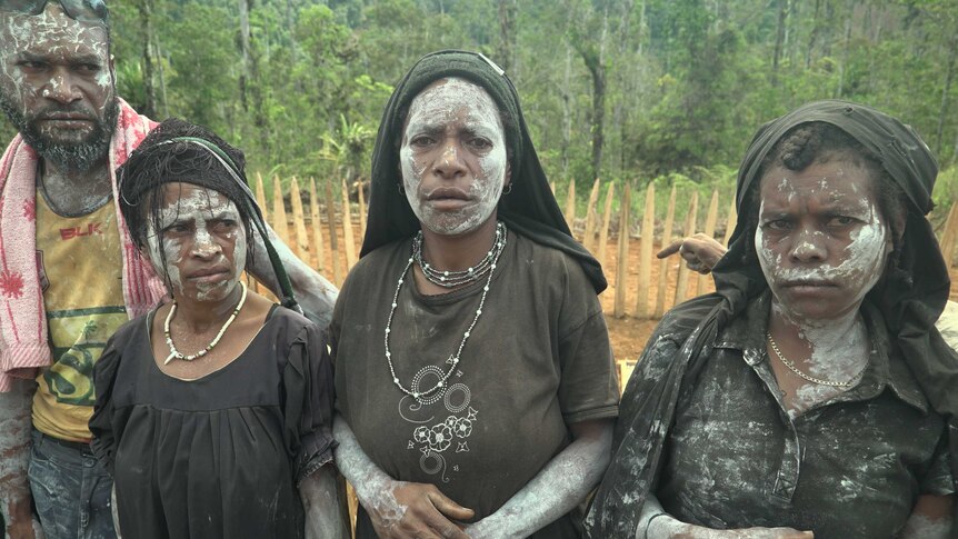 Three women and one man stand in a row with their faces painted white, mourning their lost relatives