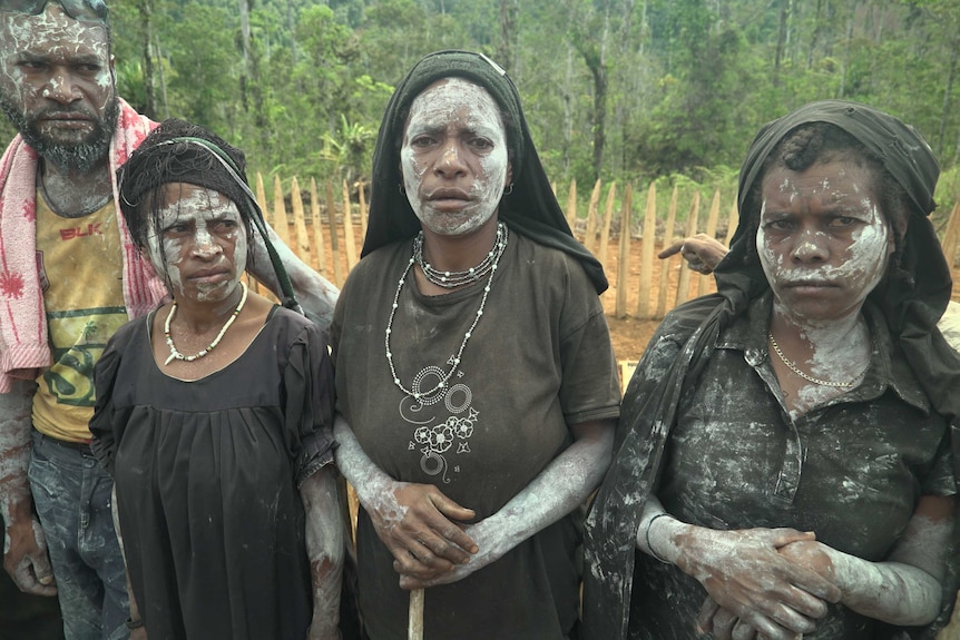 Three women and one man stand in a row with their faces painted white, mourning their lost relatives