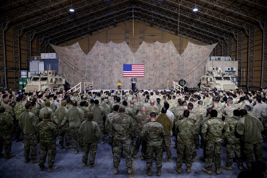 A wide-shot of an air hangar shows a crowd of US troops in camo listening to President Donald Trump.