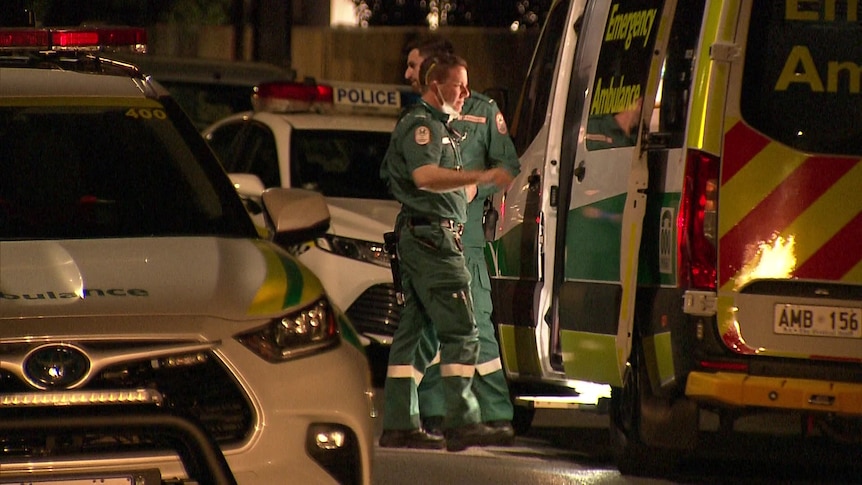 Two ambulance workers in green uniforms standing next to police cars and an ambulance.