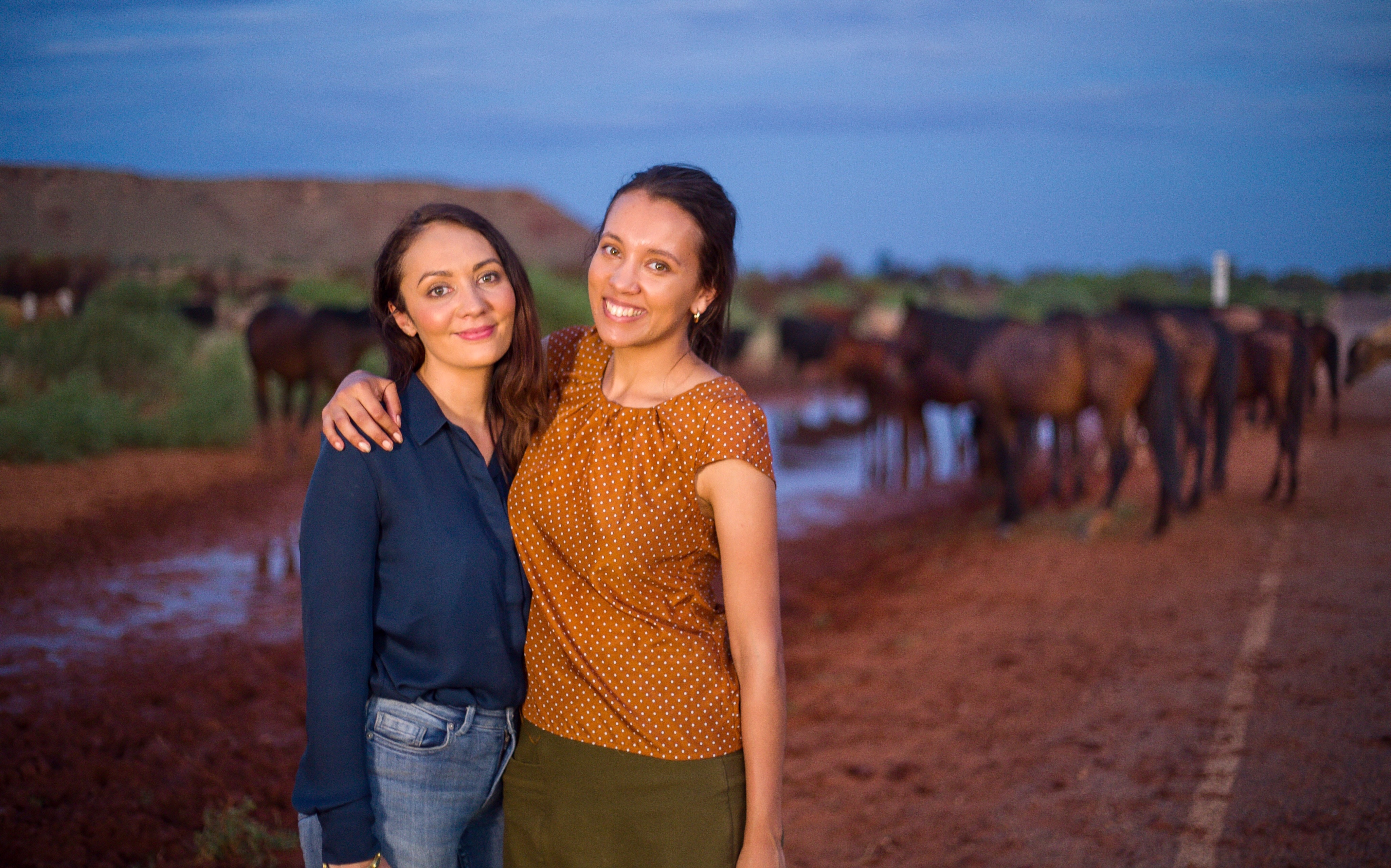 Two women with arms around each other standing in front of a pack of horses drinking at a waterhole.