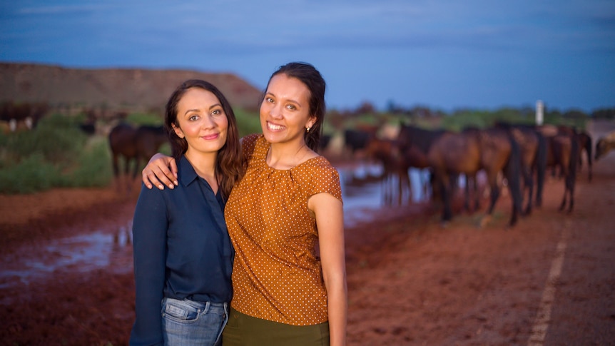 Two women with arms around each other standing in front of a pack of horses drinking at a waterhole.