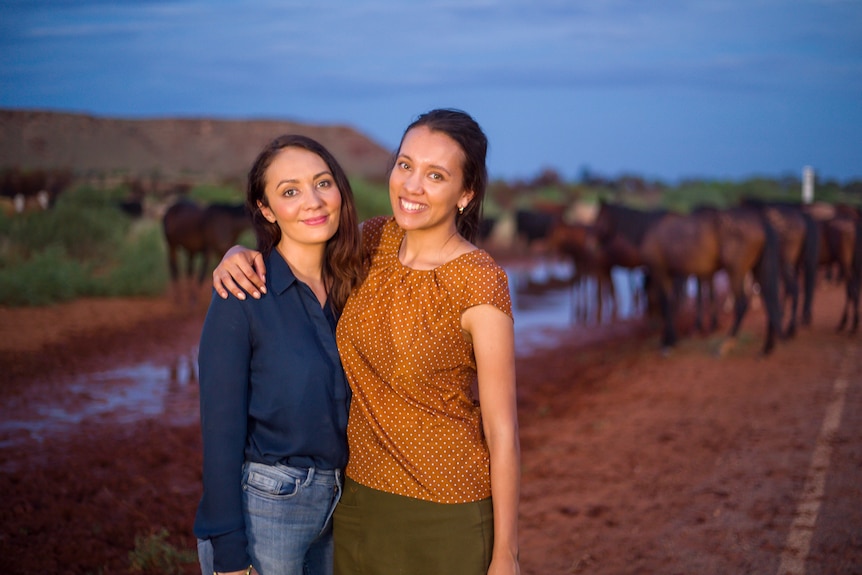 Two women with arms around each other standing in front of a pack of horses drinking at a waterhole.